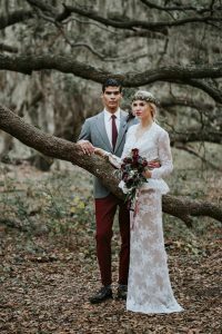 A man wearing a gray tuxedo and maroon pants and a woman wearing lacy wedding dress on a forest