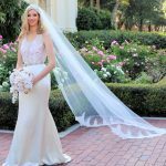 An elegant bride wearing a couture wedding dress stands gracefully in front of a vibrant flower garden, her delicate veil gently flowing in the breeze.
