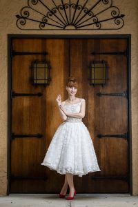Jackie, wearing a Short Wedding Dress Jackie/ Made to Order Bridal Gown, standing in front of a wooden door.