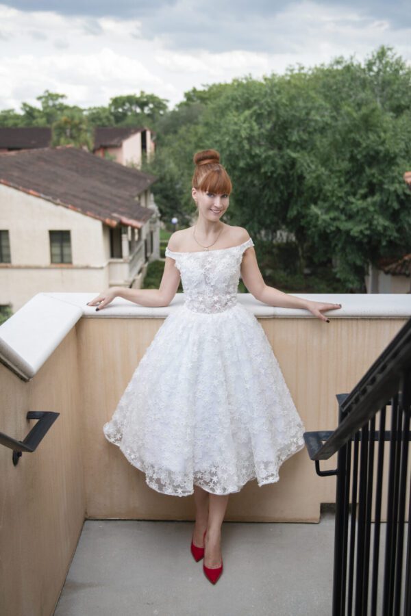 Jackie, a woman in a Short Wedding Dress Jackie/Made to Order Bridal Gown, standing on a balcony.