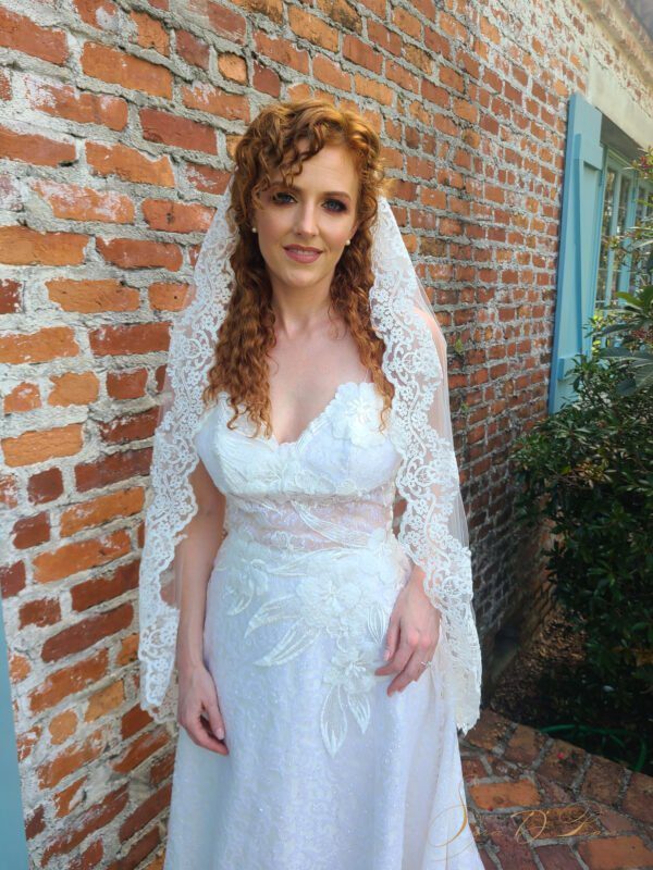 A woman in a wedding dress is standing in front of a brick wall, wearing a bridal veil.