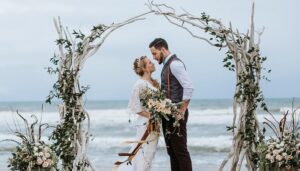 A bride and groom share a romantic kiss under a driftwood arch on the beach during their beautiful beach wedding.