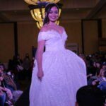 A woman in a white wedding dress struts confidently down the runway at a Bridal Fashion show.