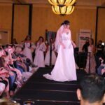 A woman in a bridal gown elegantly struts down the runway at a fashion show.