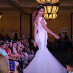 At a bridal fashion show, a woman in a white dress gracefully walks down the runway, capturing everyone's attention.