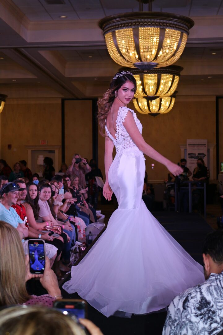 At a bridal fashion show, a woman in a white dress gracefully walks down the runway, capturing everyone's attention.