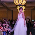 A woman in a wedding dress stuns the audience at a Bridal Fashion show as she gracefully walks down the runway.